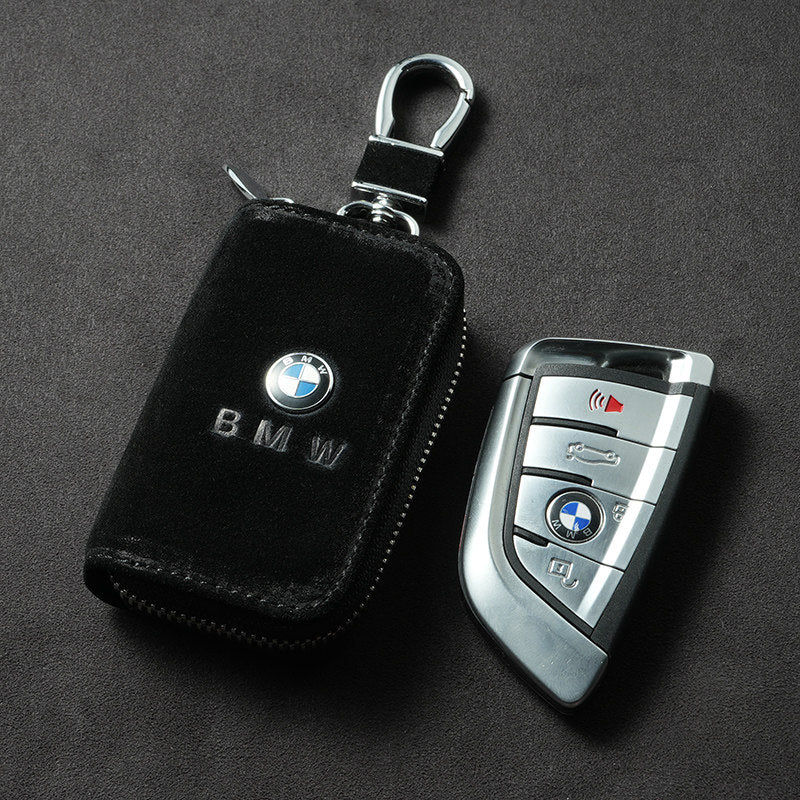 A black leather zippered car key fob cover with a silver BMW logo and keychain clip, shown next to a silver BMW key fob on a dark textured background.